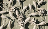 Decorative white metal or wrought iron artwork features intricate floral and leaf designs. The textured green wooden background contrasts with the light color of the metal, highlighting the delicate flower petals and swirling leaf patterns.