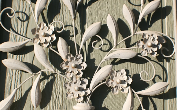 Decorative white metal or wrought iron artwork features intricate floral and leaf designs. The textured green wooden background contrasts with the light color of the metal, highlighting the delicate flower petals and swirling leaf patterns.