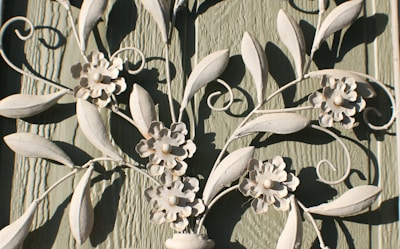 Decorative white metal or wrought iron artwork features intricate floral and leaf designs. The textured green wooden background contrasts with the light color of the metal, highlighting the delicate flower petals and swirling leaf patterns.