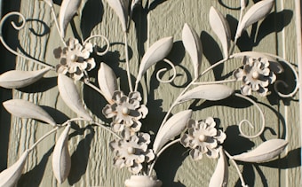 Decorative white metal or wrought iron artwork features intricate floral and leaf designs. The textured green wooden background contrasts with the light color of the metal, highlighting the delicate flower petals and swirling leaf patterns.