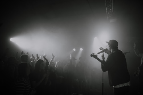 A black and white concert scene with a performer on stage holding a microphone and guitar. A crowd of people with raised hands is visible, enjoying the music under bright stage lights that create a smoky atmosphere.