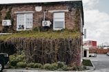 A brick building partially covered with green ivy and plants, with two air conditioning units on the wall. The windows have bars over them, and there is a small sign that reads 'GREENGO'. The scene includes a parking lot with a black vehicle on the left and more buildings in the background.