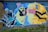 A vibrant piece of street art depicting a surreal scene where a character with a ghostly face and black hair is intertwined with a figure holding a can of spray paint. There are abstract geometric patterns and a broken heart symbol in the upper left corner.