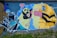 A vibrant piece of street art depicting a surreal scene where a character with a ghostly face and black hair is intertwined with a figure holding a can of spray paint. There are abstract geometric patterns and a broken heart symbol in the upper left corner.