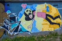 A vibrant piece of street art depicting a surreal scene where a character with a ghostly face and black hair is intertwined with a figure holding a can of spray paint. There are abstract geometric patterns and a broken heart symbol in the upper left corner.