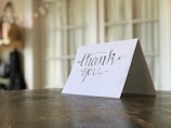 A thank you card with elegant cursive handwriting is propped up on a wooden table surface. The background is softly blurred, with a hint of warm lighting and some indistinct shapes.
