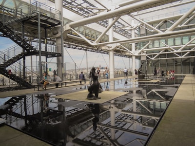 A modern architectural setting featuring an open terrace with large reflective black surfaces and a metallic structural framework. People are seated and standing around the terrace, with some looking at sculptures positioned on the reflective platforms. A metal staircase and railings are visible on the left, leading to an enclosed walkway supported by cylindrical columns.