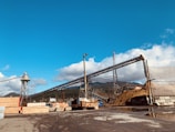 A large industrial site with several conveyor belts, piles of materials, and stacks of wooden planks. The setting is outdoors under a clear, blue sky with scattered clouds in the background. Trees and hills are visible in the distance, suggesting a rural or semi-industrial location.