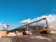 A large industrial site with several conveyor belts, piles of materials, and stacks of wooden planks. The setting is outdoors under a clear, blue sky with scattered clouds in the background. Trees and hills are visible in the distance, suggesting a rural or semi-industrial location.