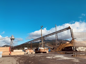 A large industrial site with several conveyor belts, piles of materials, and stacks of wooden planks. The setting is outdoors under a clear, blue sky with scattered clouds in the background. Trees and hills are visible in the distance, suggesting a rural or semi-industrial location.