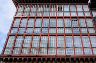 A facade of a building featuring multiple rows of large panes of glass framed in wooden grids. The structure has a reddish-brown wooden framework that creates a geometric pattern.