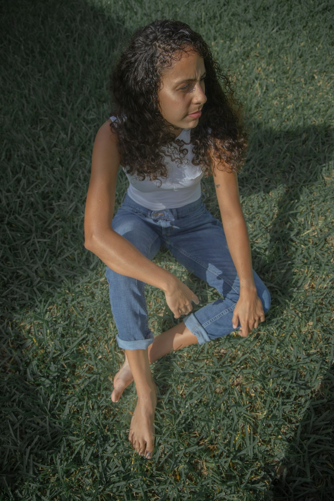 woman in white tank top and blue denim jeans sitting on green grass during daytime