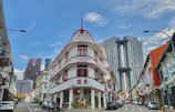 An urban street scene with a prominent white and red colonial-style building labeled 'Potato Head' at the intersection. Modern skyscrapers, including an architectural building with connecting sky gardens, are visible in the background. The street is lined with cars and colorful shophouses, and a clear blue sky with some clouds adds to the vividness of the scene.