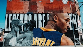 A brightly colored mural occupies a brick wall, depicting a large image of a basketball player in a jersey with the name 'Bryant' in bold letters. To the left, a touching scene shows a man and a young girl in grayscale embracing. The background of the mural reads 'LEGENDS ARE...'. The setting appears urban, with visible street signs and the walls of nearby buildings.