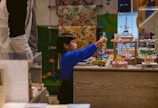 A young child wearing a blue top interacts with mechanical toys displayed on a wooden counter. The background features colorful floral and green patterns, and there are other shoppers or people nearby.