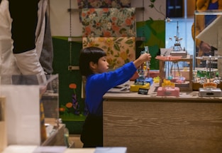 A young child wearing a blue top interacts with mechanical toys displayed on a wooden counter. The background features colorful floral and green patterns, and there are other shoppers or people nearby.