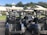 A group of golf carts is parked in a neat row outside a building with a white roof. The area is surrounded by lush greenery, including palm trees and other foliage. The sky is blue with a few clouds, creating a vibrant and inviting atmosphere.