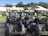 A group of golf carts is parked in a neat row outside a building with a white roof. The area is surrounded by lush greenery, including palm trees and other foliage. The sky is blue with a few clouds, creating a vibrant and inviting atmosphere.
