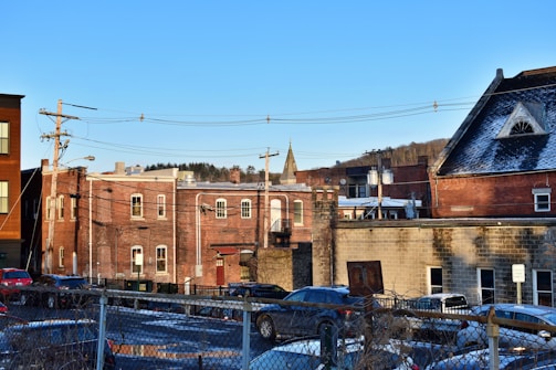 A small town scene with several brick buildings, some of which have snow on the rooftops. There are power lines and utility poles visible in the foreground. A number of parked cars are in a lot enclosed by a chain-link fence. A church spire is visible in the distance with trees and a clear blue sky in the background.
