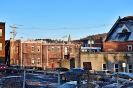 A small town scene with several brick buildings, some of which have snow on the rooftops. There are power lines and utility poles visible in the foreground. A number of parked cars are in a lot enclosed by a chain-link fence. A church spire is visible in the distance with trees and a clear blue sky in the background.
