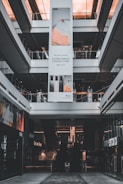 An indoor view of a shopping mall featuring a large vertical advertisement for Huawei in the center. The ad showcases mobile devices and is suspended from the ceiling. There are escalators visible beneath the ad leading to the upper floors. The architecture is modern with glass railings and neutral colors.