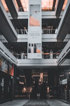 An indoor view of a shopping mall featuring a large vertical advertisement for Huawei in the center. The ad showcases mobile devices and is suspended from the ceiling. There are escalators visible beneath the ad leading to the upper floors. The architecture is modern with glass railings and neutral colors.