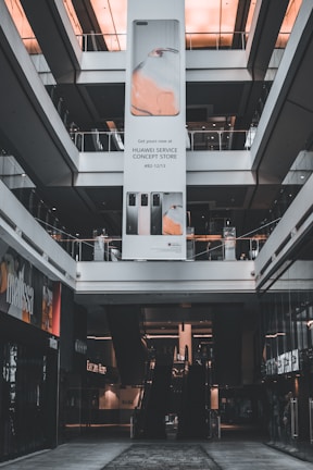 An indoor view of a shopping mall featuring a large vertical advertisement for Huawei in the center. The ad showcases mobile devices and is suspended from the ceiling. There are escalators visible beneath the ad leading to the upper floors. The architecture is modern with glass railings and neutral colors.