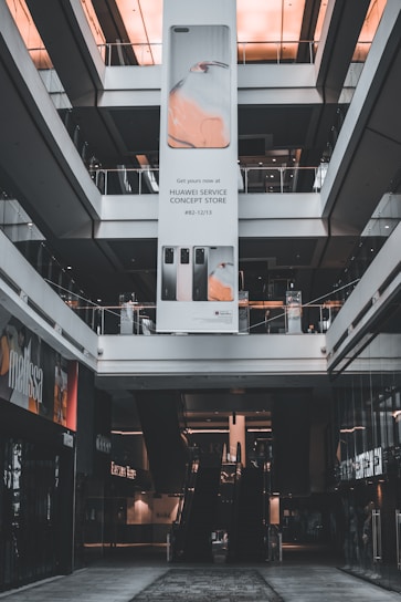 An indoor view of a shopping mall featuring a large vertical advertisement for Huawei in the center. The ad showcases mobile devices and is suspended from the ceiling. There are escalators visible beneath the ad leading to the upper floors. The architecture is modern with glass railings and neutral colors.