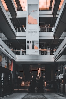 An indoor view of a shopping mall featuring a large vertical advertisement for Huawei in the center. The ad showcases mobile devices and is suspended from the ceiling. There are escalators visible beneath the ad leading to the upper floors. The architecture is modern with glass railings and neutral colors.