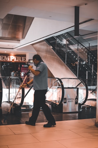 A person cleaning the floor with a mop in a shopping mall near an escalator. The surroundings include a sign for 'General Waste' and advertisements for various shops.