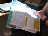 A person is reading a newsletter or brochure while seated. The document contains information related to property management, with the title 'Northshore Property Management' and a notable 'Ray White' advertisement. The setting includes a wooden table, a file, and cardboard boxes in the background.