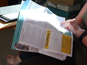 A person is reading a newsletter or brochure while seated. The document contains information related to property management, with the title 'Northshore Property Management' and a notable 'Ray White' advertisement. The setting includes a wooden table, a file, and cardboard boxes in the background.