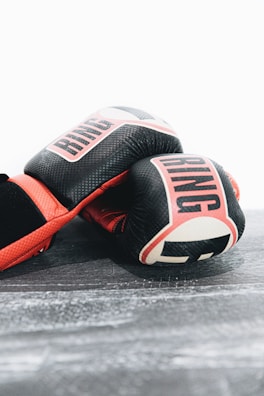A pair of boxing gloves with a black and red color scheme resting on a textured surface. The gloves have the word 'RING' printed in white letters with a bold font.