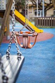 A playground scene features a focus on a children's swing seat with orange handles and metal chains. In the background, there is a yellow slide attached to a climbing structure. The ground is covered in a blue, red, and green rubber safety mat, surrounded by wooden fencing.
