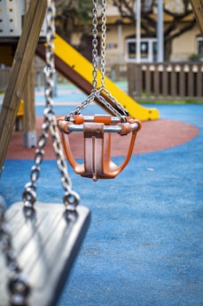 A playground scene features a focus on a children's swing seat with orange handles and metal chains. In the background, there is a yellow slide attached to a climbing structure. The ground is covered in a blue, red, and green rubber safety mat, surrounded by wooden fencing.