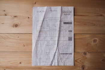 A document is laid out on a wooden surface. It appears to be a printed or photocopied sheet with tables and text written in Arabic. The paper is slightly crumpled and shows signs of wear. The wooden surface has a natural finish with visible knots and grain patterns.