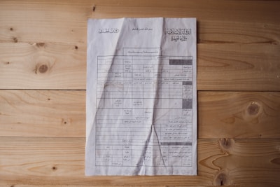 A document is laid out on a wooden surface. It appears to be a printed or photocopied sheet with tables and text written in Arabic. The paper is slightly crumpled and shows signs of wear. The wooden surface has a natural finish with visible knots and grain patterns.