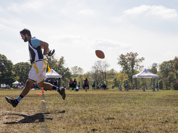 A person is engaged in a sports activity, running on a grassy field. The person is wearing a sports jersey and shorts. They are positioned in mid-air as they run, and a football is visible in front of them. In the background, there are several people along with tents and trees, indicating an outdoor sports event.