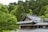 A traditional Japanese building with a gray tiled roof nestled among lush green trees. The architecture features intricate wooden details, typical of classic Japanese design, with large windows allowing for natural light. The surrounding forest is dense and vibrant, contributing to a serene, natural atmosphere.