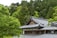 A traditional Japanese building with a gray tiled roof nestled among lush green trees. The architecture features intricate wooden details, typical of classic Japanese design, with large windows allowing for natural light. The surrounding forest is dense and vibrant, contributing to a serene, natural atmosphere.