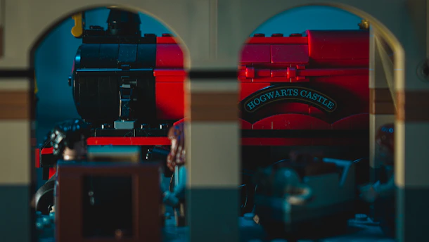 A red model train with the sign 'Hogwarts Castle' is visible through an archway. The scene appears to be constructed from LEGO bricks, featuring detailed elements such as windows and wheels. The lighting creates a subtle, moody atmosphere.