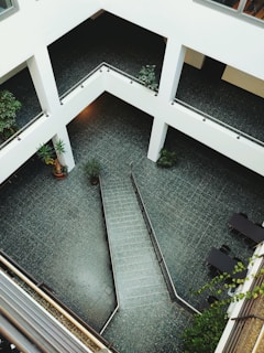 A modern architectural space with a multi-level design featuring open corridors and a central atrium area. The floor is tiled with gray or dark-colored tiles, and the surroundings include various green potted plants. Staircases and railings add structure to the open area. The overall design is minimalist with a focus on clean lines and open spaces.