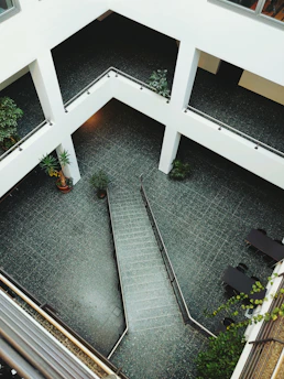 A modern architectural space with a multi-level design featuring open corridors and a central atrium area. The floor is tiled with gray or dark-colored tiles, and the surroundings include various green potted plants. Staircases and railings add structure to the open area. The overall design is minimalist with a focus on clean lines and open spaces.