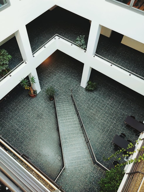 A modern architectural space with a multi-level design featuring open corridors and a central atrium area. The floor is tiled with gray or dark-colored tiles, and the surroundings include various green potted plants. Staircases and railings add structure to the open area. The overall design is minimalist with a focus on clean lines and open spaces.