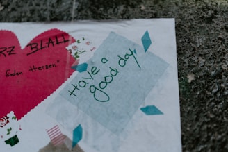 A collage featuring a large red heart with handwritten text in black, along with a smaller blue sticky note displaying the phrase 'have a good day' in green cursive writing. The background consists of a speckled, textured stone surface.