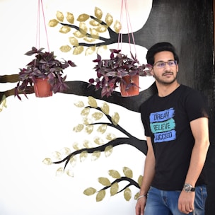 A person wearing glasses and a black T-shirt with motivational words stands beside a wall mural. The mural features branches with gold leaves and two hanging red potted plants. The person is looking slightly to the side, exuding a relaxed demeanor.