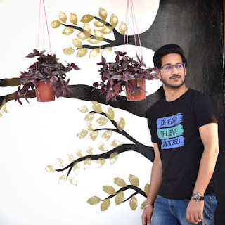 A person wearing glasses and a black T-shirt with motivational words stands beside a wall mural. The mural features branches with gold leaves and two hanging red potted plants. The person is looking slightly to the side, exuding a relaxed demeanor.