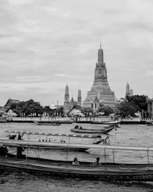 A black and white photograph of a large, intricately designed temple tower situated by a river. Several longboats are visible on the water, with people seated inside. The temple, adorned with detailed patterns, features multiple spires and is surrounded by trees and smaller buildings.