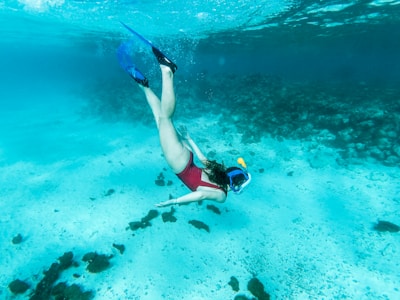 A person is diving underwater wearing a snorkel and fins. The clear blue water reveals a sandy sea floor scattered with dark rocks and small patches of coral. The person is facing downwards, gracefully extending their arms and legs in a relaxed, exploratory pose.