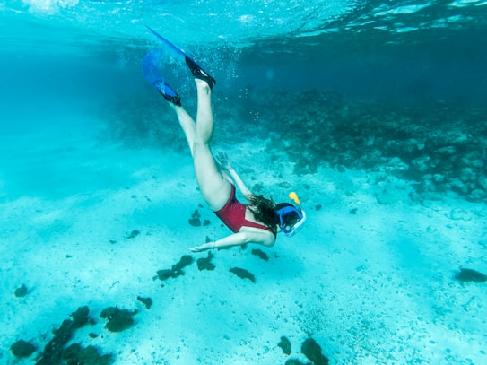 A person is diving underwater wearing a snorkel and fins. The clear blue water reveals a sandy sea floor scattered with dark rocks and small patches of coral. The person is facing downwards, gracefully extending their arms and legs in a relaxed, exploratory pose.